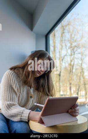 Junge Frau in Strickpullover und Brille zeichnen oder arbeiten auf Tablet-pc Stockfoto