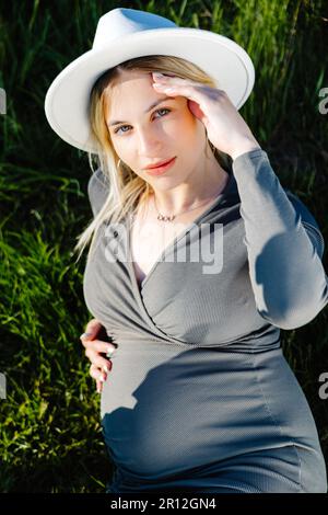 Schöne schwangere Frau mit Hut und Kleid, die auf dem Gras liegt. Frau, die ein Baby erwartet Stockfoto