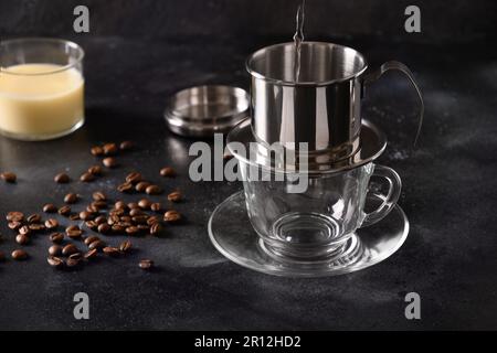 Gießen Sie heißes Wasser in den Filter, um vietnamesischen Kaffee mit süßer Kondensmilch in einer Glasbecher auf schwarzem Hintergrund zuzubereiten. Schließen. Stockfoto