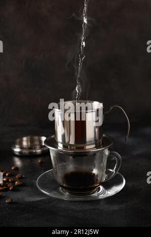 Gießen Sie heißes Wasser in den Filter, um vietnamesischen Kaffee in einem Glasbecher auf schwarzem Hintergrund zuzubereiten. Schließen. Stockfoto