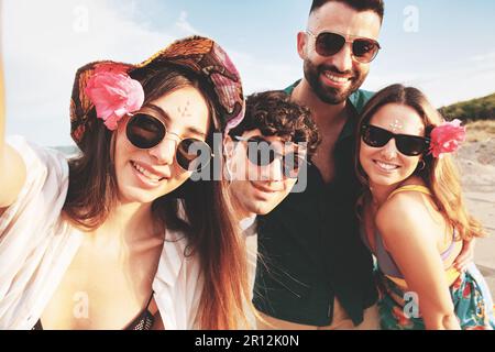 Eine Gruppe von vier glücklichen jungen kaukasischen Freunden, zwei Männern und zwei Frauen, machen an einem sonnigen Sommertag ein Selfie an einem Sandstrand, lächeln und genießen Stockfoto