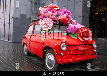 Retro-Auto mit großen Blumen in der Nähe des Einkaufszentrums in der Stadt. Ein altes rotes Auto an einem bewölkten Tag. Stockfoto