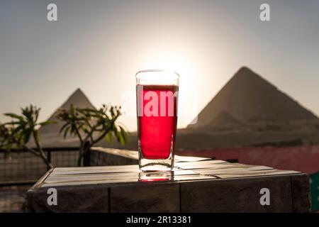 Traditionelle arabische Kräutertee-Karkade in einem Glas vor dem Hintergrund der Großen Pyramiden von Gizeh. Schöne Aussicht auf die Attraktion im Restaurant Stockfoto