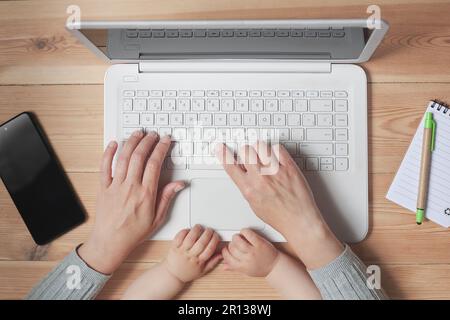 Ansicht eines Laptops von oben nach unten, wobei die Hände einer Frau und eines Babys sichtbar sind. Die Frau arbeitet, während das Baby spielt. Stockfoto