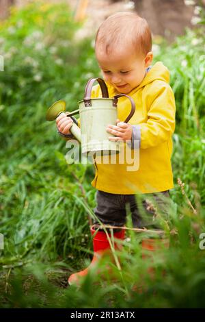 Ein kleiner Junge mit gelbem Regenmantel und Gummistiefeln steht in einer Pfütze inmitten einer Wiese, hält einen Stock und schaut auf das Wasser. Stockfoto