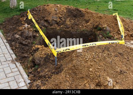 Eine quadratische Grube, die in den Boden gegraben wurde, neben dem öffentlichen Bürgersteig und mit einem gelben Band eingezäunt, als Zeichen der Gefahr. Kommunaler Service funktioniert, Proble eliminieren Stockfoto