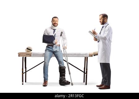 Mann mit Halskragen, orthopädischem Stiefel und Armschlinge sitzt auf einem Therapietisch und Arzt schreibt ein isoliertes Dokument auf weißem Hintergrund Stockfoto