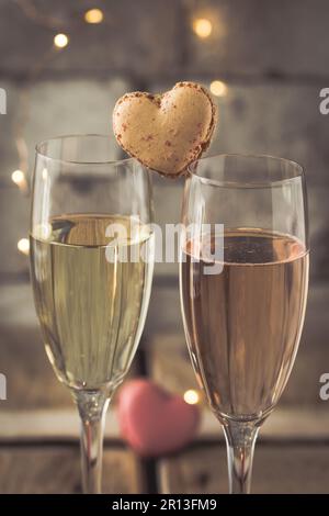 Zwei Gläser mit Sekt und herzförmigen Makronen auf einem rustikalen Holztisch, romantische Atmosphäre, vertikal Stockfoto