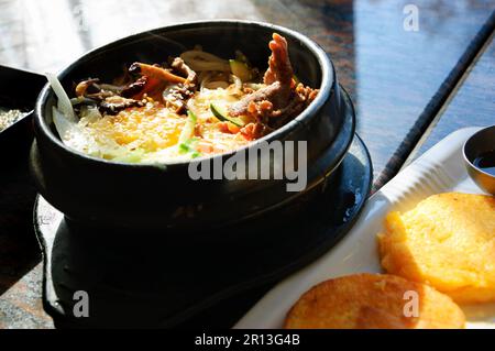 Traditionelles koreanisches Gericht. Bibimbap in einer beheizten Steinschale und gebratenen Sojabohnengaletten. Stockfoto