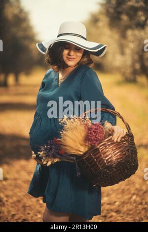 Eine schwangere Frau mit einem blauen Kleid, einem weißen Hut und einem Basketballkorb mit trockenen Blumen in einem Olivenfeld mit geringer Schärfentiefe. Stockfoto