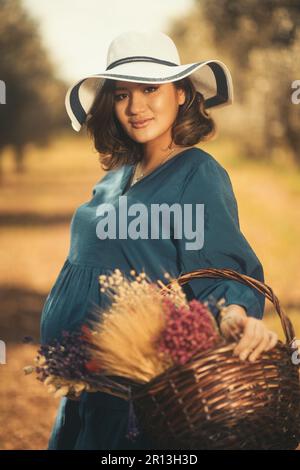Eine schwangere Frau mit einem blauen Kleid, einem weißen Hut und einem Basketballkorb mit trockenen Blumen in einem Olivenfeld mit geringer Schärfentiefe. Stockfoto