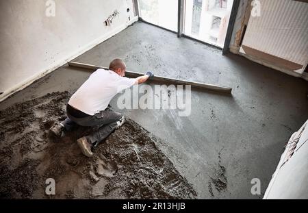 Draufsicht eines Bauarbeiters, der die Estriche auf dem mit Sandzement-Mischung bedeckten Boden platziert. Mann glättet und nivelliert die Oberfläche mit geradem Schneidmesser, während der Boden in der Wohnung gehoben wird. Stockfoto
