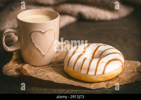 Donut auf einem dunklen Holztisch, Kaffeetasse mit Herz und Wollpullover im Hintergrund, gemütliche Atmosphäre, Kaffeepause, vertikal Stockfoto