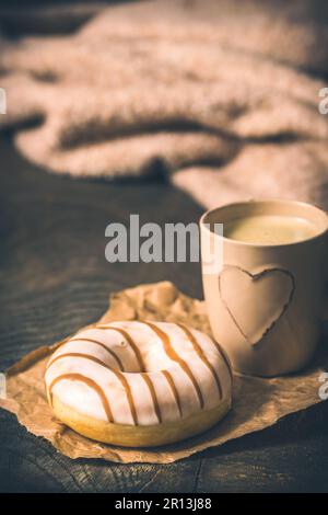 Donut auf einem dunklen Holztisch, Kaffeetasse mit Herz und Wollpullover im Hintergrund, gemütliche Atmosphäre, Kaffeepause, vertikal Stockfoto