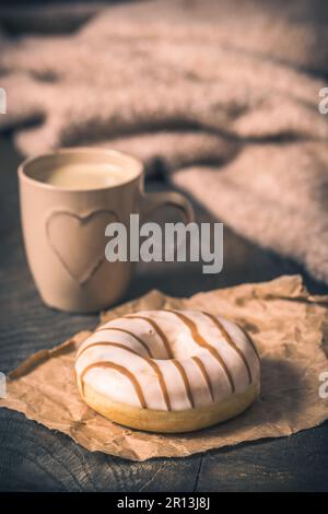 Donut auf einem dunklen Holztisch, Kaffeetasse mit Herz und Wollpullover im Hintergrund, gemütliche Atmosphäre, Kaffeepause, vertikal Stockfoto