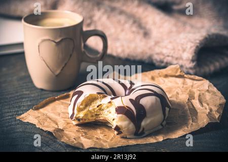 Donut auf einem dunklen Holztisch, Kaffeetasse mit Herz und Wollpullover im Hintergrund, gemütliche Atmosphäre, Kaffeepause Stockfoto