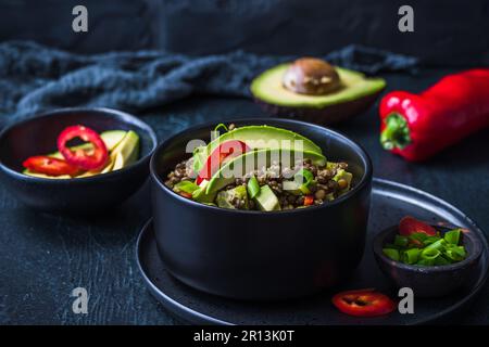 Linsensalat mit Avocado und rotem Pfeffer in einer schwarzen Schüssel auf schwarzem Hintergrund. Vegetarische und vegane Speisen. Stockfoto