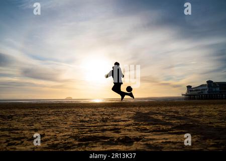 Silhouette eines kleinen Jungen, der bei Sonnenuntergang am Strand in Weston-super-Mare, Somerset, Großbritannien, Fußball spielt. Stockfoto