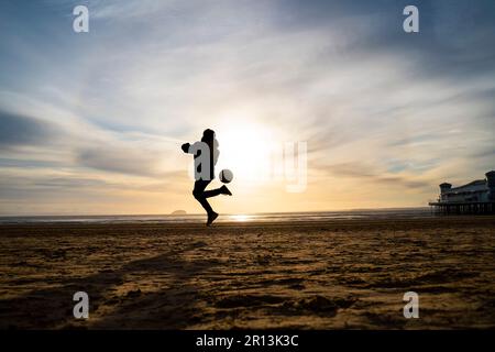 Silhouette eines kleinen Jungen, der bei Sonnenuntergang am Strand in Weston-super-Mare, Somerset, Großbritannien, Fußball spielt. Stockfoto