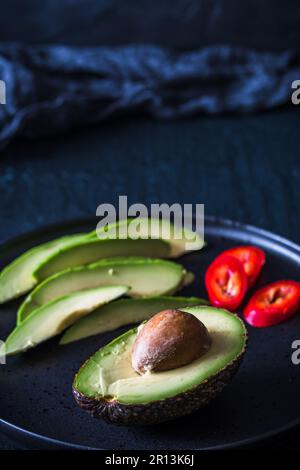 Halbe Avocado mit Grube und Avocadoscheiben auf schwarzer Platte, vertikal mit Kopierraum Stockfoto