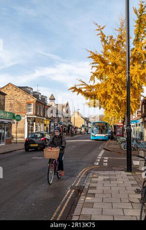 Herbst am Mill Road Broadway, Cambridge, Großbritannien. Ein Bus hält an einer Bushaltestelle neben einer Parade kleiner unabhängiger Geschäfte mit einem Fahrradfahrer im Vordergrund. Stockfoto