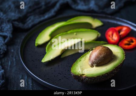 Halber Avocado mit Grube und Avocado-Scheiben auf schwarzem Teller Stockfoto