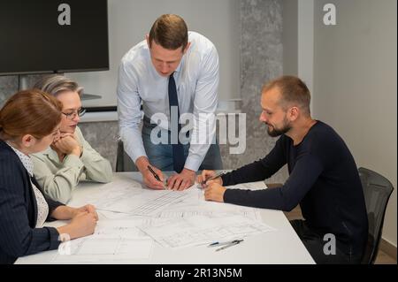 Ein weißer Mann steht und ändert eine Zeichnung, drei Kollegen sitzen an einem Tisch und hören ihm zu. Stockfoto