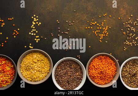 Mehrfarbige Linsen in Schüsseln auf braunem Hintergrund, verstreut gelb und braun, grüne und orangefarbene Linsen, gesunde Hülsenfrüchte, Draufsicht Stockfoto