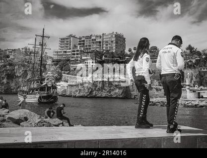 Ein Polizist und eine Polizistin stehen am Rande des alten Hafens in der Altstadt von Kaleici, Antalya, Türkei (Turkiye). Dahinter entspannen sich die Leute im Stockfoto