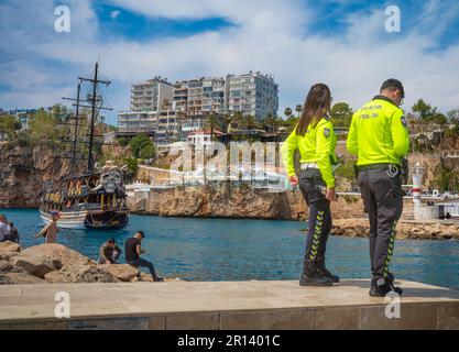 Ein Polizist und eine Polizistin stehen am Rande des alten Hafens in der Altstadt von Kaleici, Antalya, Türkei (Turkiye). Dahinter entspannen sich die Leute im Stockfoto