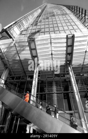 Leute, die die Rolltreppe nach The Shard in London, England, hochfahren. Balck und Weiß mit farbigem Burst auf gut sichtbarer Jacke eines Werkers. Stockfoto