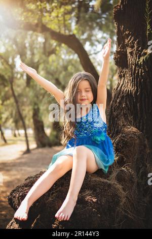 Wunderschönes glückliches Mädchen, das im Park mit blauem Kleid ohne Schuhe spielt, auf dem Baum sitzt und vor Glück schreit, während die Familie Kinder genießt Stockfoto