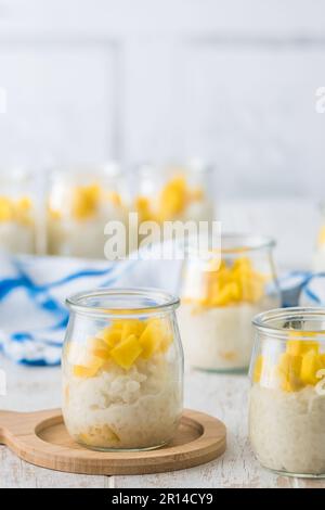 Kokosreis-Pudding mit Mango in Dessertgläsern auf weißem Holzhintergrund, vertikal Stockfoto