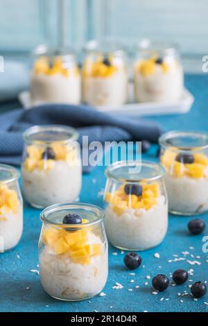 Kokosreis-Pudding mit Mango in Dessertgläsern auf blauem Hintergrund, vertikal Stockfoto