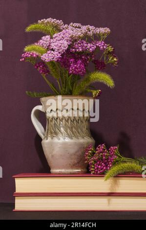 Stilleben mit violetten Schafgartenblumen in traditioneller Keramikvase auf Büchern Stockfoto