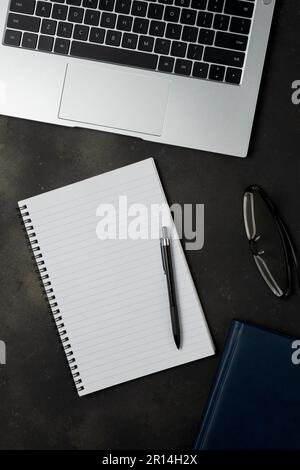 Flach liegender Schreibtisch mit Blick von oben. Arbeitsplatz mit leeren Notizbüchern, Laptops, Bleistiften und Brillen auf schwarzem Hintergrund. Stockfoto