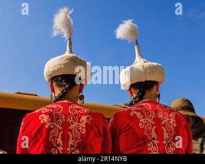 Rückansicht von zwei kirgisischen Damen in traditionellem Kostüm mit leuchtend roten, gestickten Jacken und hohen weißen Federhüten Stockfoto