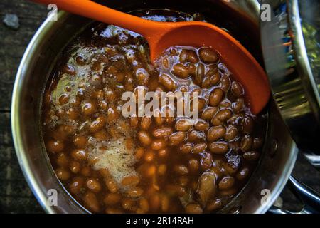Nahaufnahme von gebackenen Bohnen in einem Gericht auf dem Tisch mit einem orangefarbenen Servierlöffel, perfektes Sommergericht beim Kochen oder Treffen Stockfoto
