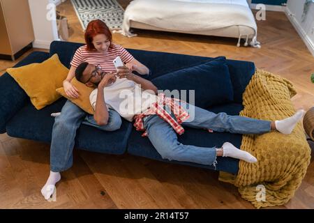 Entspanntes, vielseitiges Familienpaar-Wochenendruhe zu Hause auf der Couch, lächelnd auf dem Smartphone im Internet surfen Stockfoto
