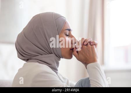 Nahaufnahme. Porträt einer jungen arabischen Frau in einem Hijab zu Hause, die ihre Hände zusammenfaltete, ihre Augen schloss und betete, an Depressionen leidet, über die Lösung von Problemen nachdenkt. Stockfoto