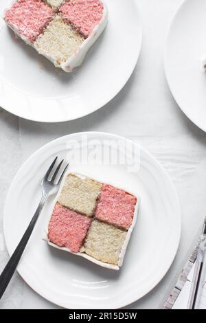 Draufsicht auf battenburger Kuchenstück auf weißem Teller, Mandel- und Erdbeerschwammkuchen bedeckt mit weißem Fondant, Querschnitt aus rosa und gelbem Karo Stockfoto