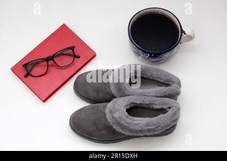 Graue flauschige, gepolsterte Hausschuhe, sehr bequem neben einem Notizbuch, Stift, Buch, Gläsern und Kaffee für einen Moment der Entspannung und Ruhe Stockfoto