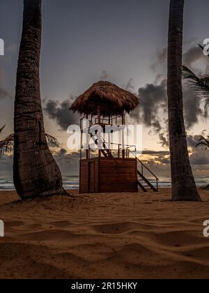 Eine Holzhütte an einem tropischen Sandstrand in der Karibik. Diese Hütte mit Strohdach liegt an der Küste der Dominikanischen Republik. Die Sonne geht auf. Stockfoto