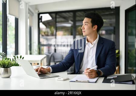 Ein gutaussehender, kluger asiatischer Geschäftsmann oder männlicher Chef sitzt an seinem Schreibtisch, schaut aus dem Fenster und träumt von seinem Geschäftserfolg. Stockfoto