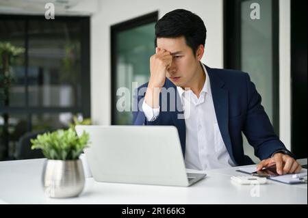 Gestresster asiatischer Geschäftsmann in formellen Business-Anzügen sitzt an seinem Schreibtisch, sieht sich seinen Laptop-Bildschirm an und denkt behutsam an einen Plan zur Lösung des Stockfoto