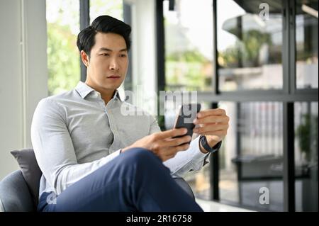 Ein attraktiver asiatischer Millennials-Geschäftsmann, der sein Smartphone nutzt und seine Nachrichten oder sozialen Medien abruft, während er sich in einer Bürolounge entspannt. Stockfoto