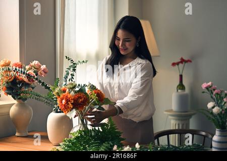 Attraktive und glückliche junge Asiatin, die ihr Wohnzimmer mit wunderschönen Vasen voller frischer Blumen dekoriert. Freizeitkonzept Stockfoto