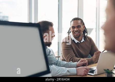 Nehmen Sie sich immer Zeit zum Lachen, auch während der Arbeit. Zwei gutaussehende Geschäftsleute sitzen im Büro und verwenden ein Notebook, während ihre Kollegen mit ihnen arbeiten Stockfoto