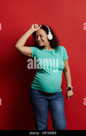 Erwachsene schwangere Frau tanzt Musik über Kopfhörer und genießt wunderbare Momente einer glücklichen, sorgenfreien Schwangerschaft Stockfoto