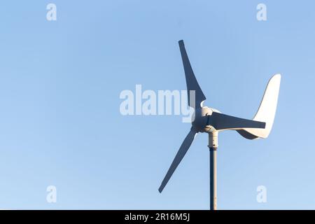 Detaillierte Aufnahme des Kopfes einer kleinen weißen Windturbine mit rotierenden Flügeln vor einem klaren blauen Himmel. Auf der linken Seite des im verfügbarer Kopierbereich Stockfoto